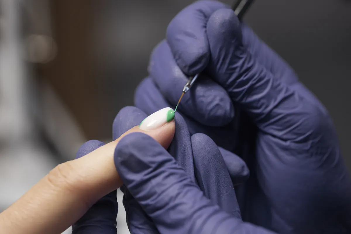 Eine Nageldesignerin mit violetten Handschuhen malt mit einem feinen Pinsel ein Detail auf einen Nagel