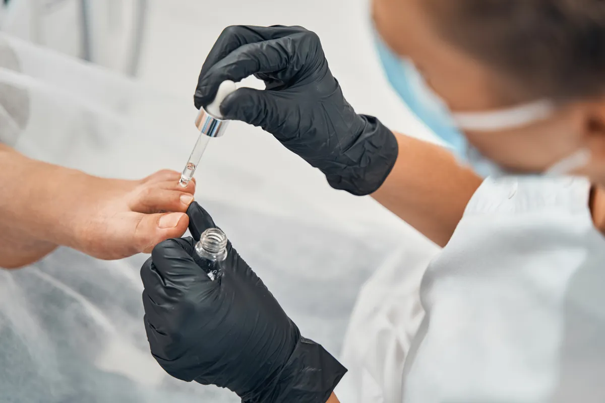 Eine Nageldesignerin mit schwarzen Handschuhen trägt mit einer Pipette Öl auf die Nägel einer Kundin auf