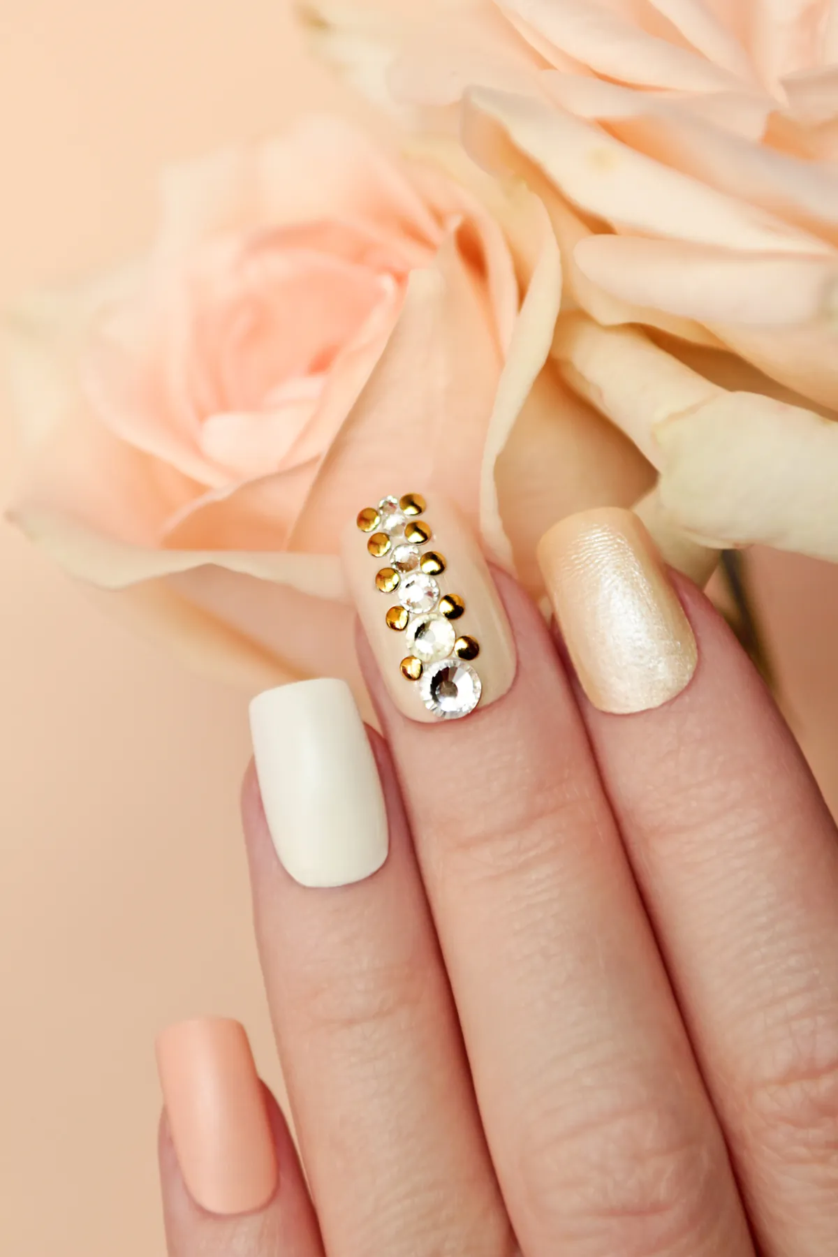 Pfirsich- und cremefarbene Nägel mit goldenen Nieten neben blassen Rosen